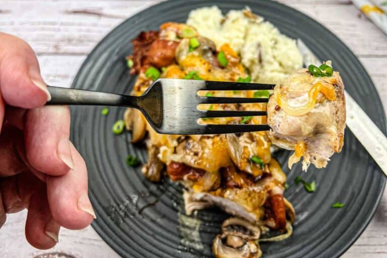 A fork holding a bite of chicken with mushrooms and cheese, over a black plate with rice and a chicken dish topped with green onions.