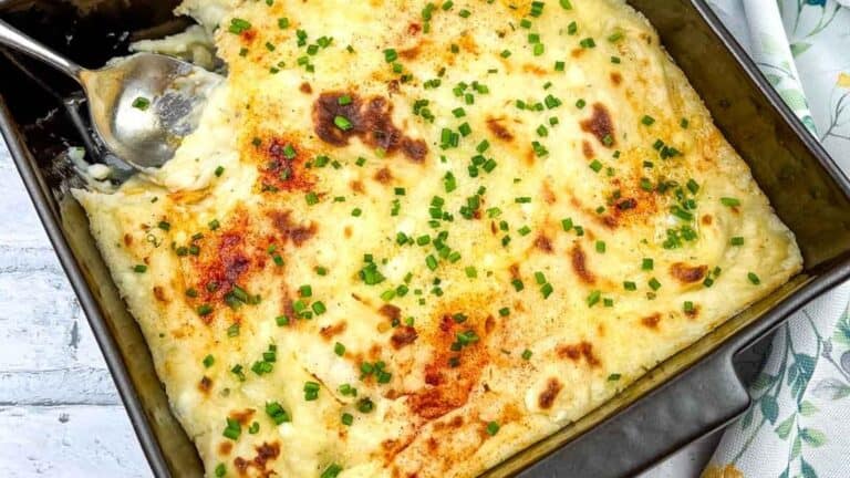 Baked mashed potatoes with browned top, garnished with chopped chives, in a rectangular baking dish with a serving spoon.