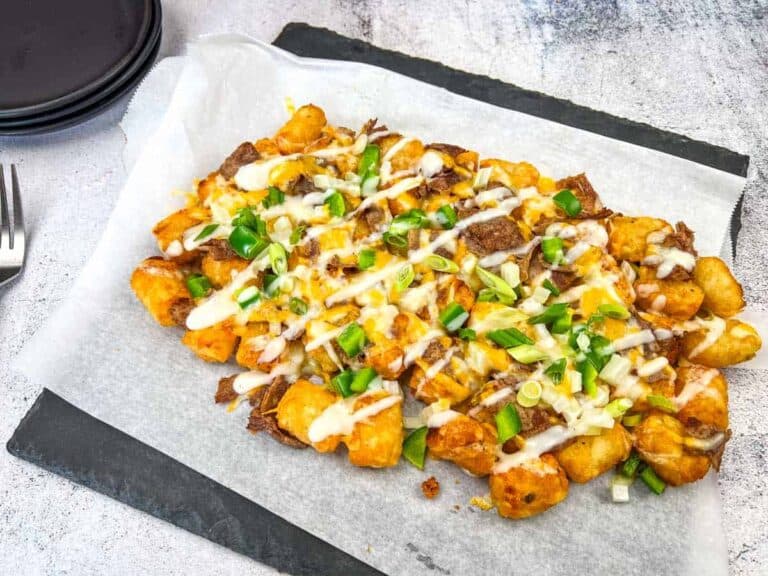 A platter of tater tots topped with melted cheese, sliced beef, chopped green onions, and drizzles of white sauce on parchment paper.