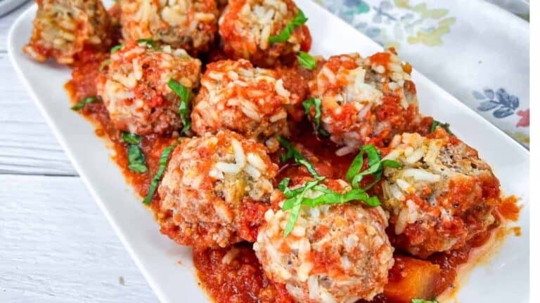 A rectangular white plate with rice and tomato-covered meatballs, garnished with chopped fresh herbs, on a wooden table.