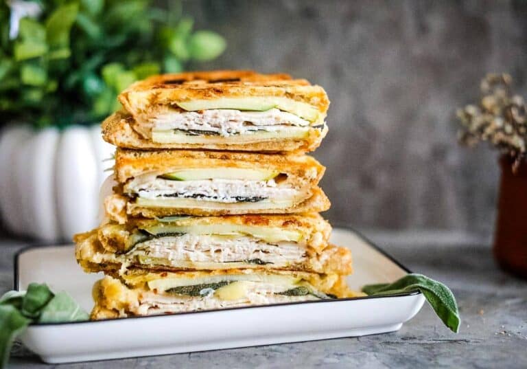 Stacked grilled cheese sandwiches with turkey, zucchini, and cheese on a white plate, placed on a gray countertop with plants in the background.