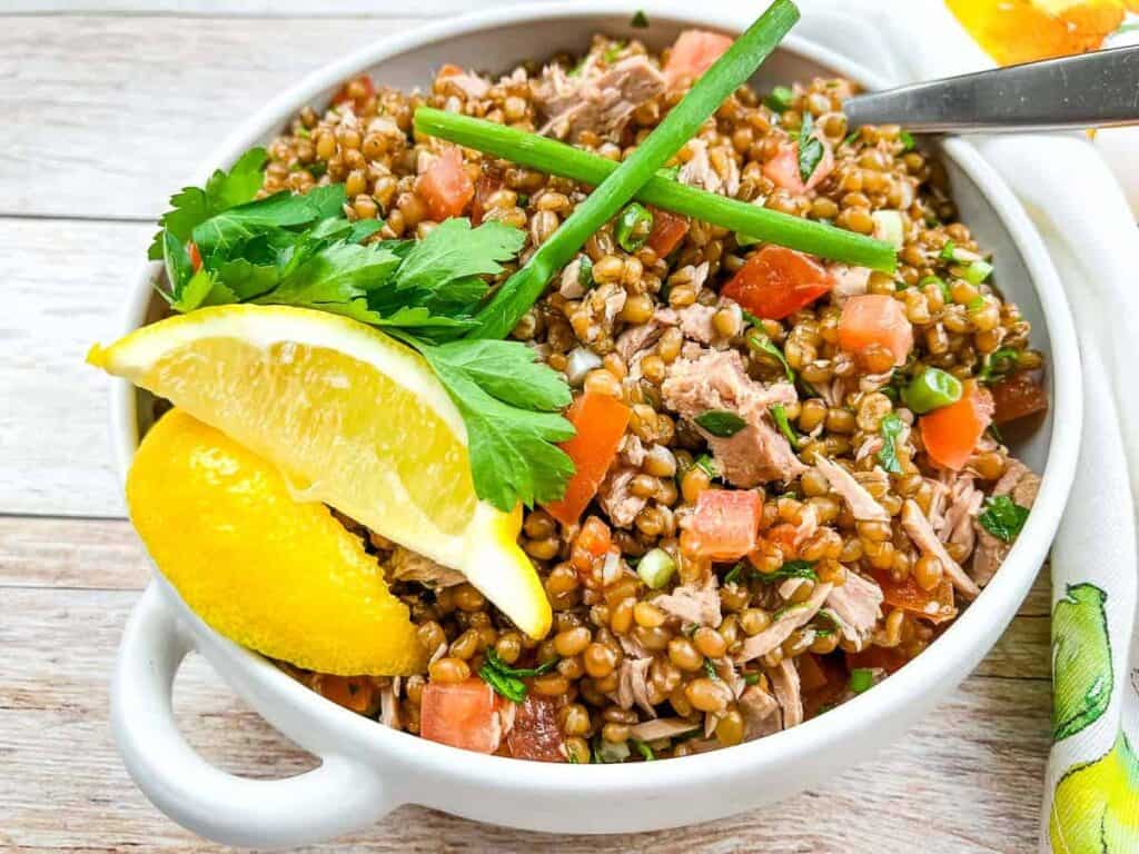 A bowl of grain salad with chopped tomatoes, green onions, and parsley, garnished with lemon wedges and fresh chives, on a wooden surface.