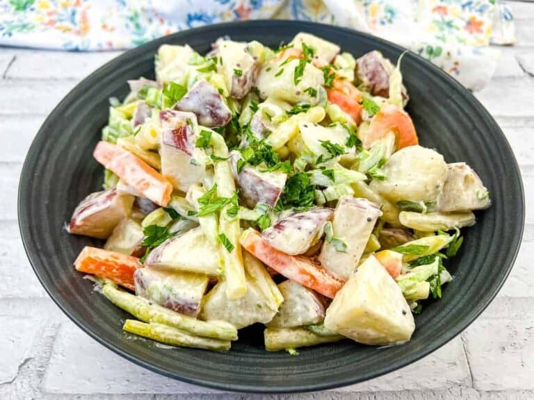 A black plate with potato salad, featuring chunks of potatoes, carrots, and green beans, garnished with chopped parsley.