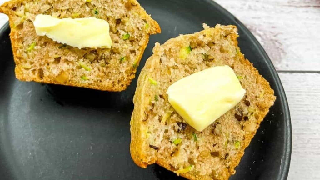 Two slices of zucchini bread with walnuts, each topped with a pat of butter, on a black plate.