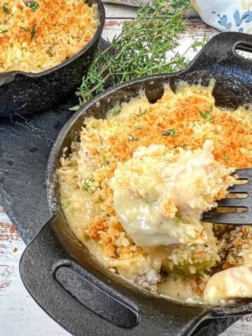 Two cast-iron skillets filled with creamy, baked casserole topped with golden breadcrumbs and garnished with fresh thyme sprigs, set on a wooden surface. A fork rests in one serving.