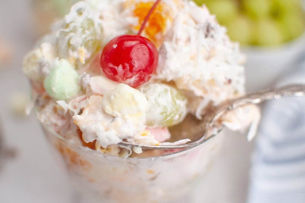 A glass of Ambrosia Salad featuring fruits, accompanied by a spoon for serving.