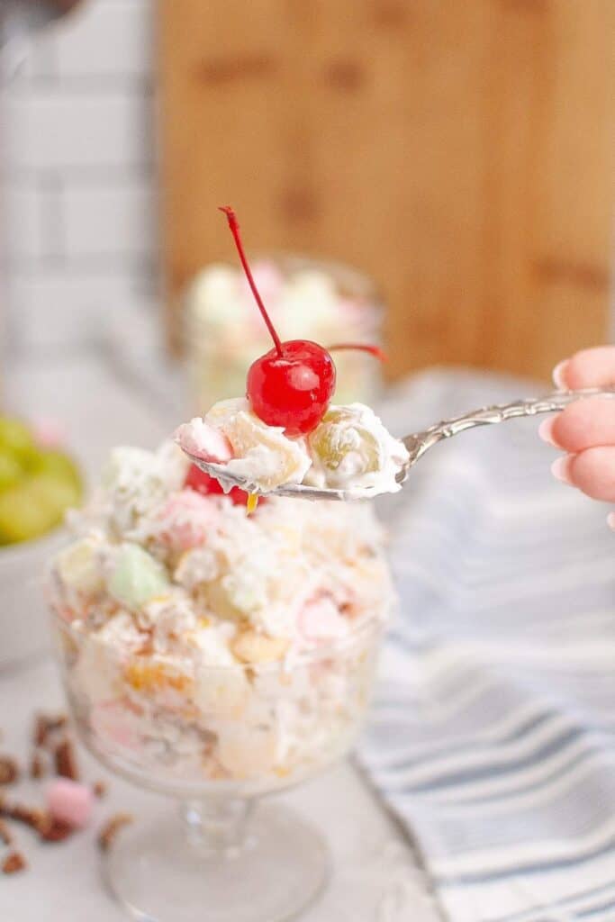 A hand holds a spoon above a bowl filled with colorful Ambrosia fruit salad.