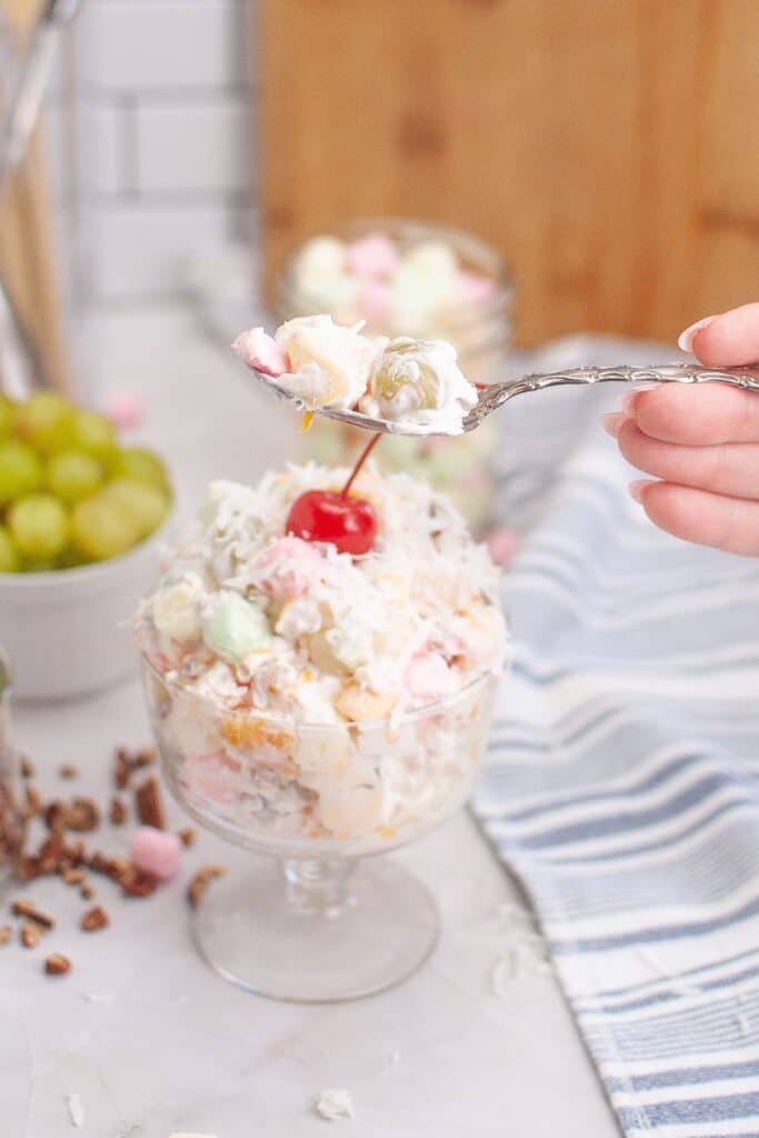 A hand holding a spoon above a bowl filled with colorful Ambrosia fruit salad.