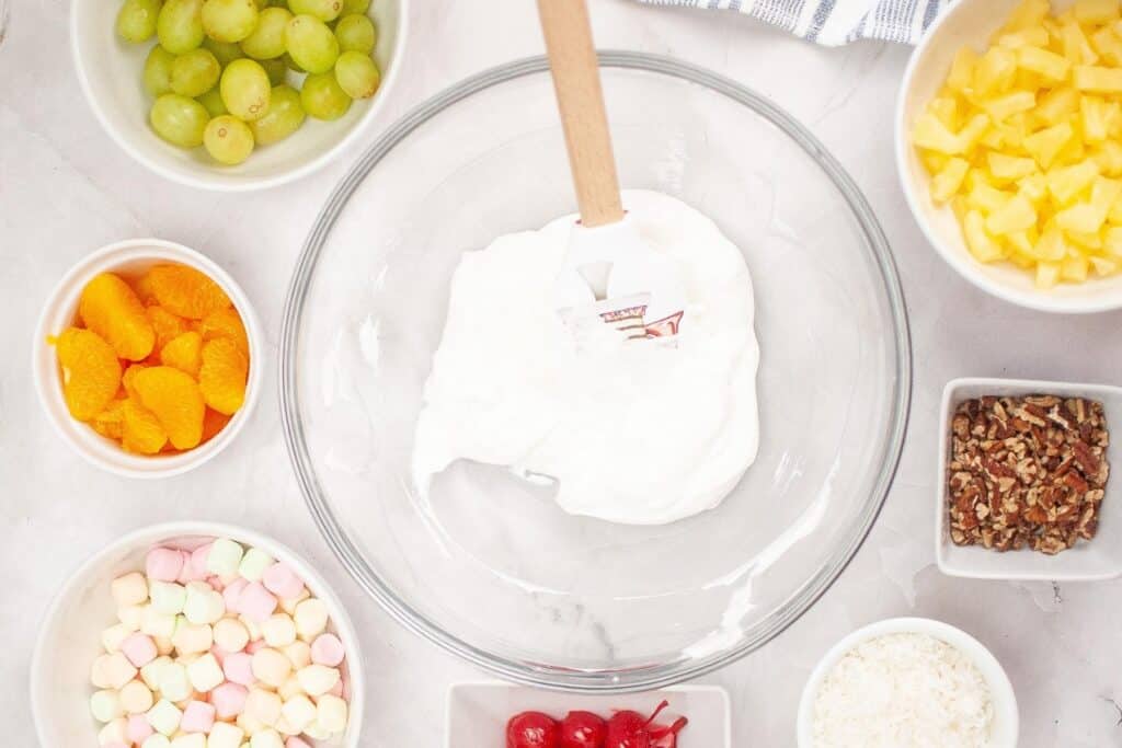 A variety of fruits and toppings displayed for an Ambrosia Salad, showcasing oranges, grapes, pineapples, cherries, marshmallows, pecans, coconut and whipped cream.