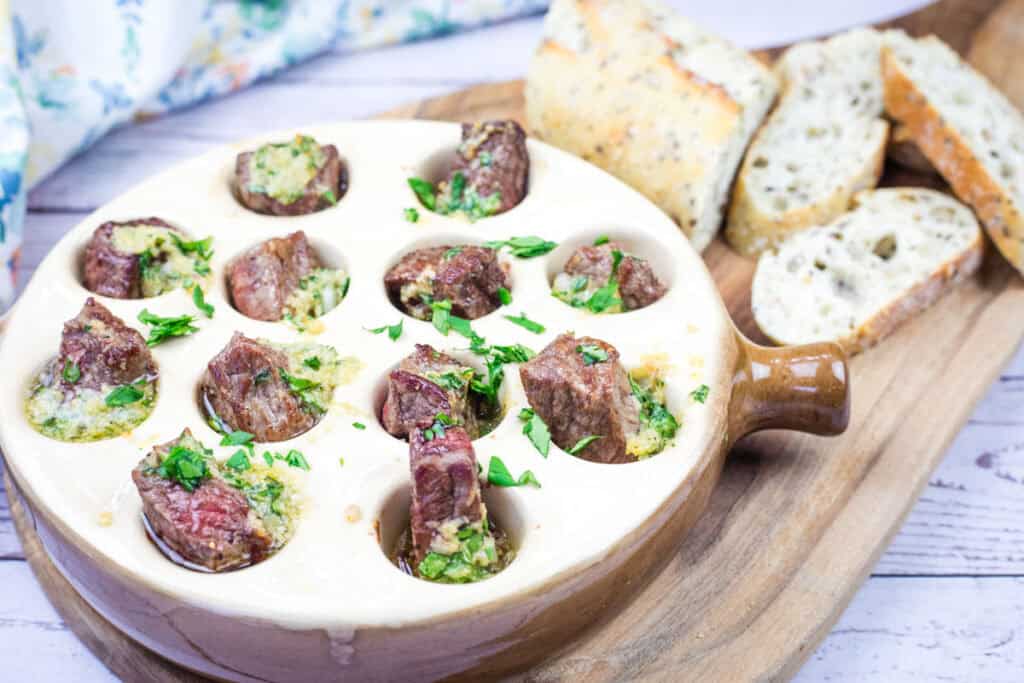 A bowl of garlic butter steak bites and bread on a wooden cutting board.