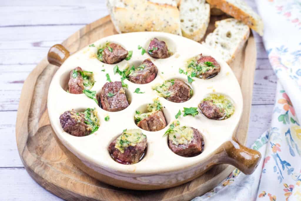 A bowl of garlic butter steak bites and bread on a wooden cutting board.
