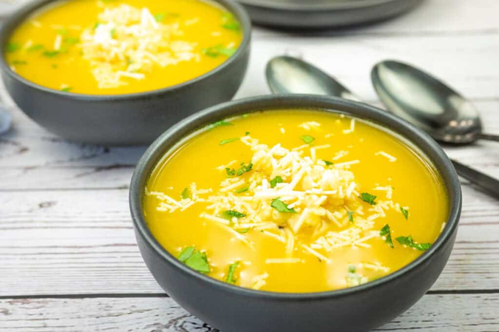 Bowls of Nonna's Pastina Soup garnished with shredded cheese and herbs on a wooden table, with spoons beside them.