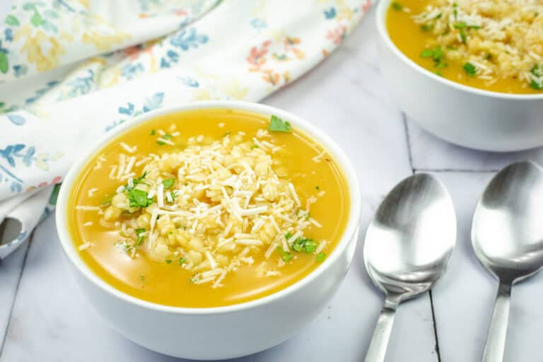 A bowl of Nonna’s pastina soup topped with shredded cheese and herbs on a marble surface.