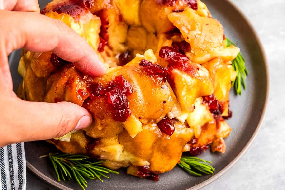 A hand pulls apart a Mini Cranberry-Brie Pull-Apart Bread garnished with rosemary, on a gray plate.