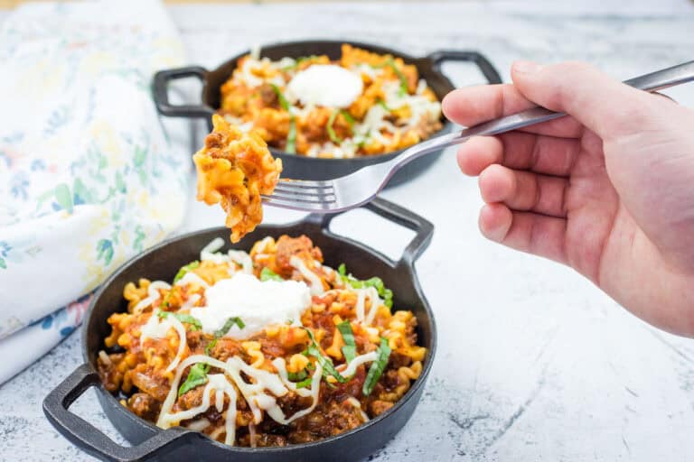 Hand holding a fork with lasagna over a mini black skillet of lasagna.