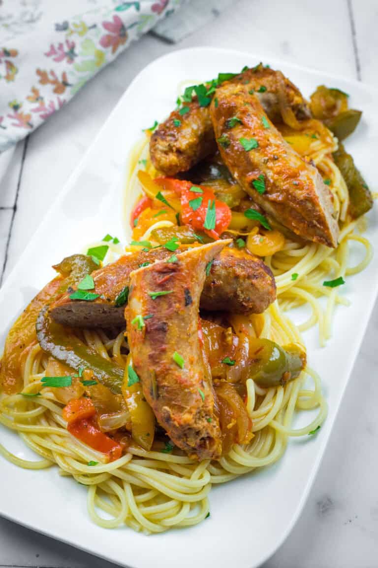 Pasta topped with grilled sausages, sliced bell peppers, and chopped herbs on a rectangular white plate.