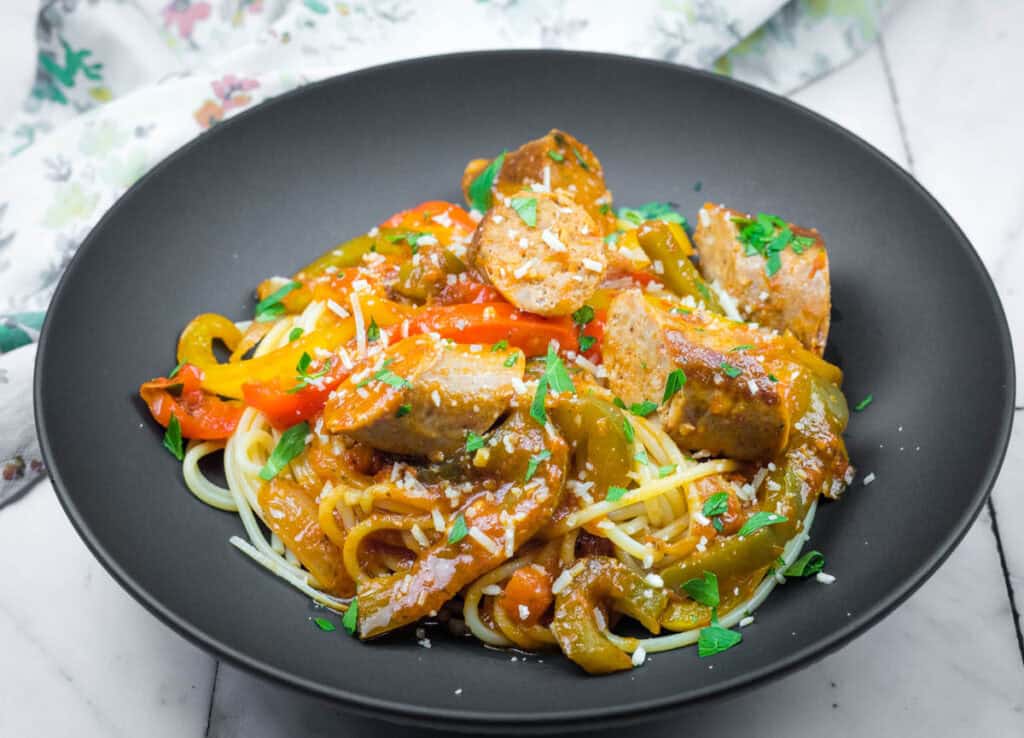 A black bowl filled with spaghetti topped with sliced sausage, red and green bell peppers, and parsley, with grated cheese sprinkled over the dish.