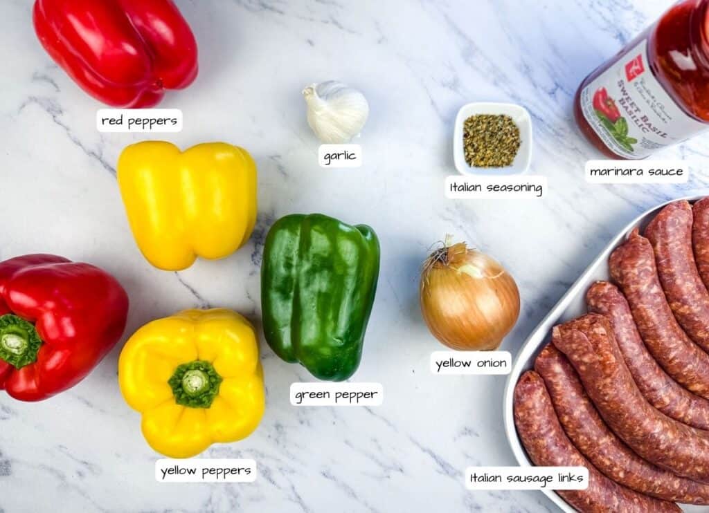 Various ingredients are laid out on a marble surface, including bell peppers, garlic, onion, Italian seasoning, marinara sauce, and Italian sausage links.