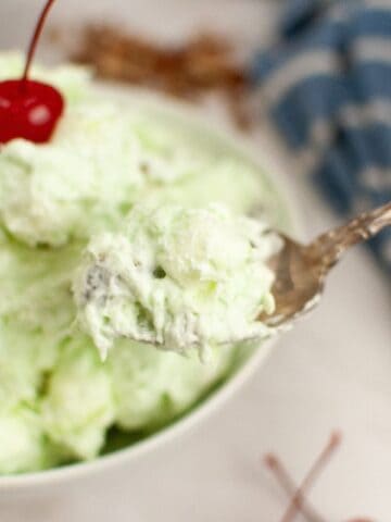 A spoonful of Watergate salad garnished with a cherry, is held over a bowl.