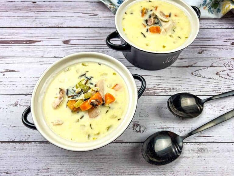 Two bowls of creamy soup with chicken, carrots, and herbs on a wooden surface, accompanied by two metal spoons.