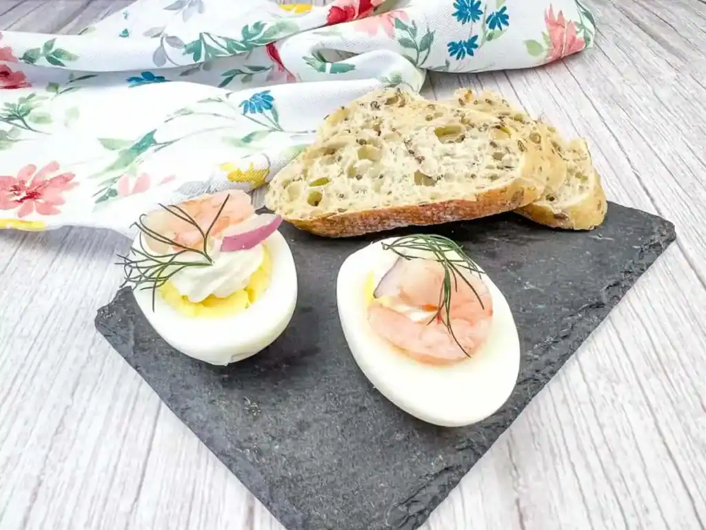 Two deviled egg halves topped with shrimp and dill on a slate plate, served with slices of multigrain bread.