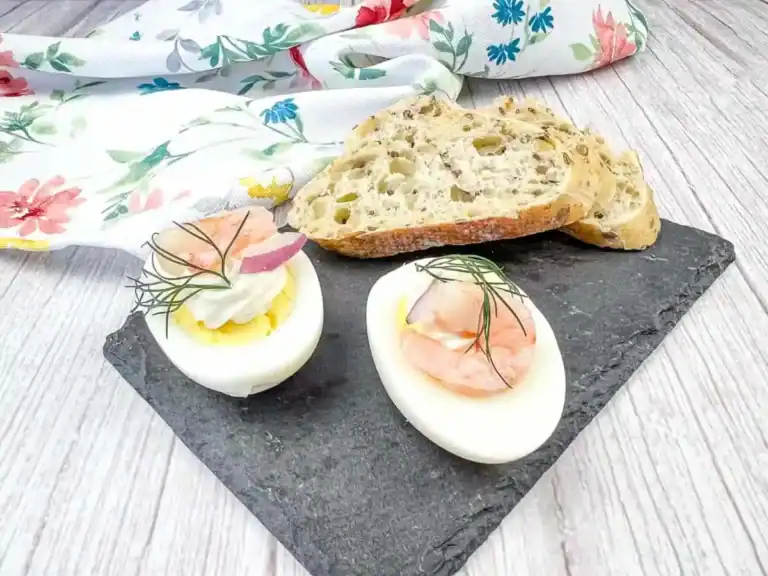 Two deviled egg halves topped with shrimp and dill on a slate plate, served with slices of multigrain bread.