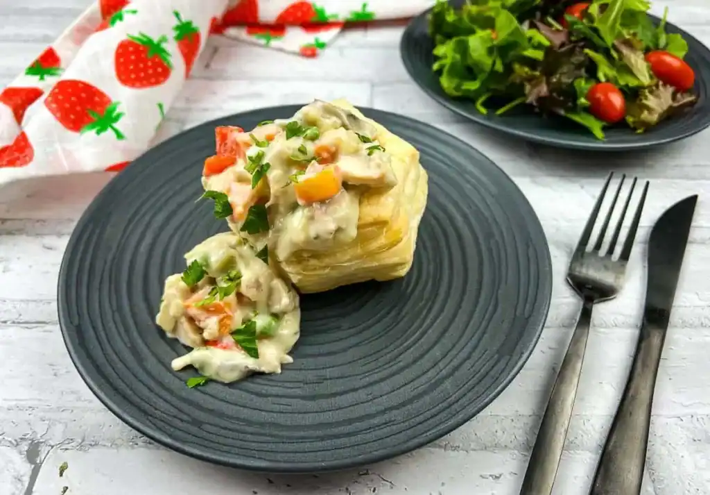 Pastry filled with creamy sauce and vegetables on a black plate, accompanied by a small salad with mixed greens and cherry tomatoes on another plate.