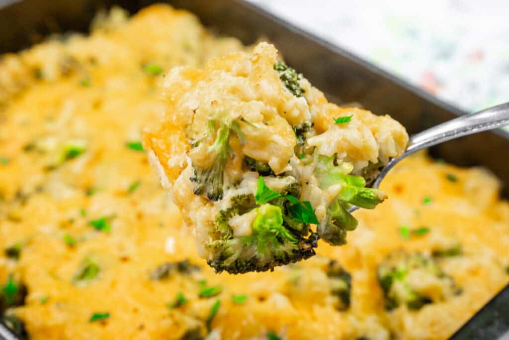 Close-up of a spoon holding a serving of cheesy broccoli and rice casserole, garnished with parsley, with the remaining dish in the background.