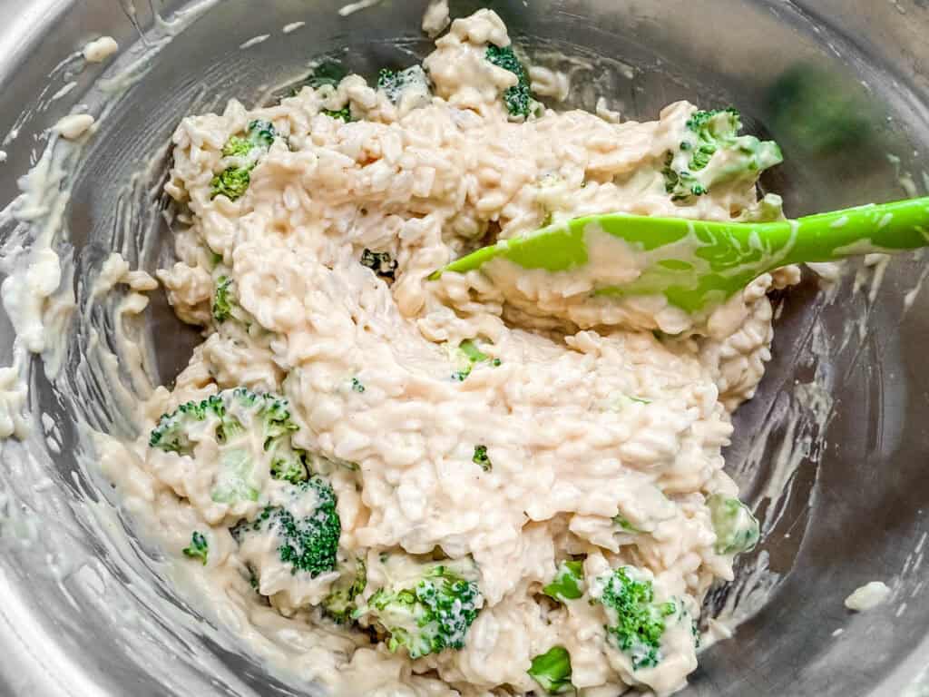 A metal bowl containing a mixture of rice, broccoli, and creamy sauce. A green spatula is visible, partially immersed in the mixture.