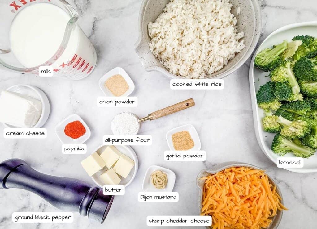 Ingredients for Broccoli Rice Casserole on a marble surface: milk, cooked white rice, broccoli, cream cheese, paprika, all-purpose flour, onion powder, garlic powder, butter, Dijon mustard, sharp cheddar cheese, black pepper.