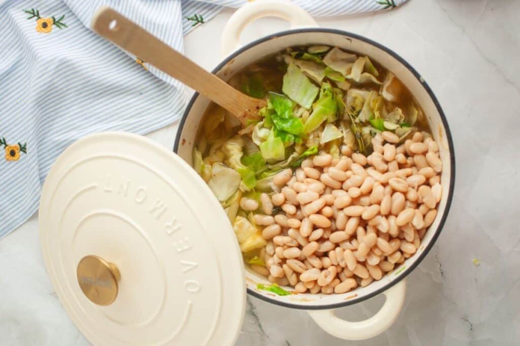 A pot of soup with white beans and cabbage, placed on a marble surface. A wooden spoon rests inside the pot. The pot lid is partially open, and a striped cloth is nearby.