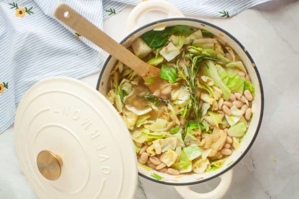 A pot containing a mix of white beans, chopped cabbage, leafy greens, and herbs. The pot is partially covered with its lid, and a wooden spoon rests inside.