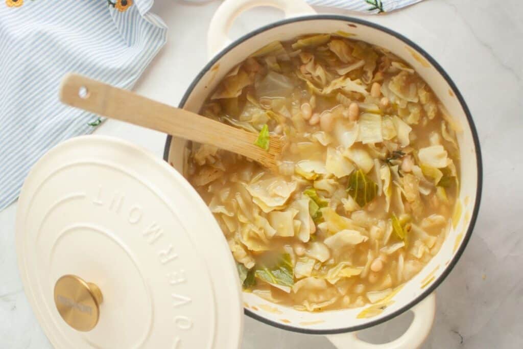 A pot of cabbage soup with white beans, featuring a wooden spoon inside. The pot is partially covered with a lid. The background includes a striped cloth with small floral accents.