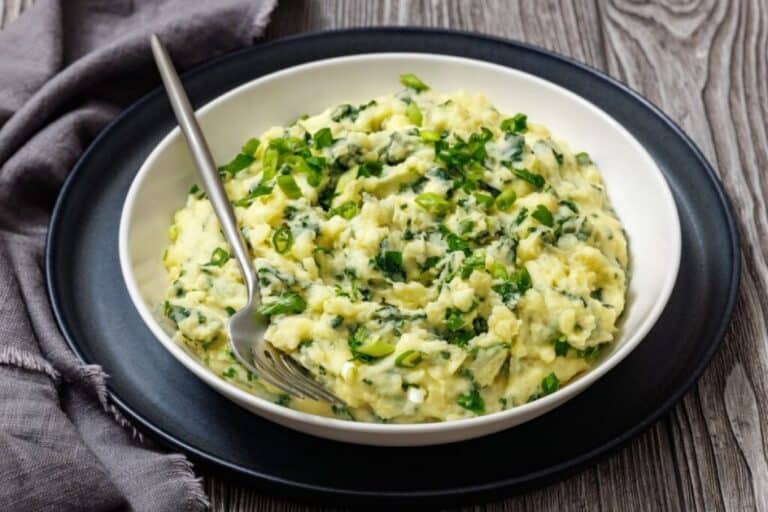 A bowl of Colcannon with Kale, placed on a black plate with a fork in the dish, with a textured wooden background and grey cloth on the side.