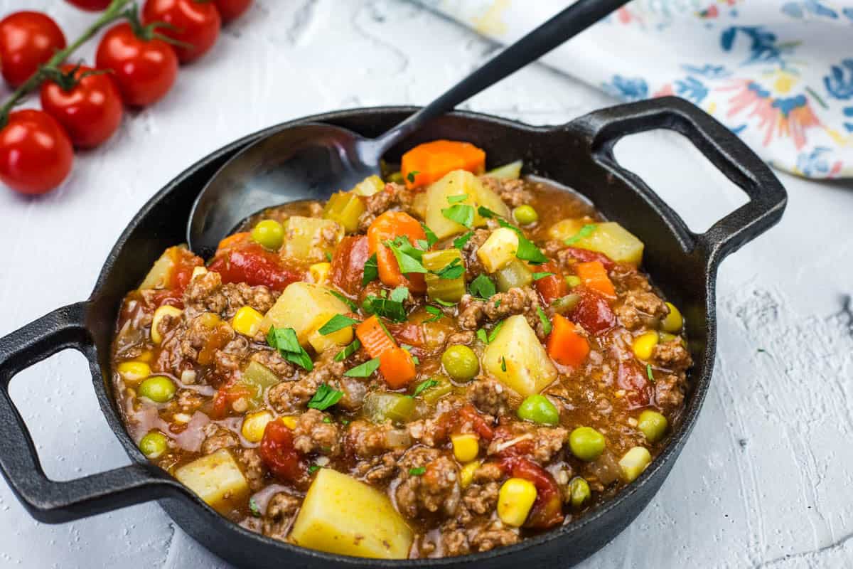 A black bowl of Mom's Hamburger Stew, featuring potatoes, peas, corn, carrots, and garnished with parsley. A spoon rests inside the bowl. Cherry tomatoes are in the background.