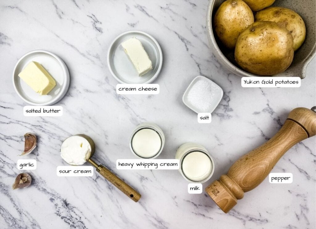 Grandma's mashed potato ingredients on a marble surface, including butter, cream cheese, sour cream, garlic, milk, salt, pepper, and a bowl of potatoes with label.