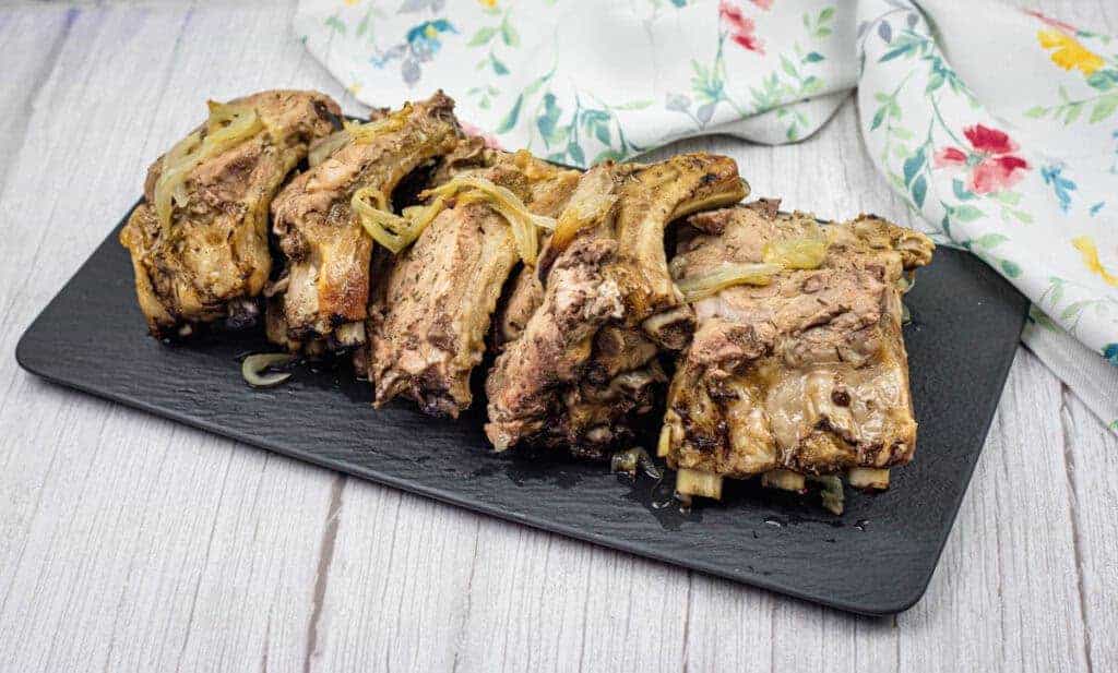 Oven Baked Ribs with cooked onions on a black rectangular slate platter, set on a wooden surface. A floral-patterned cloth is partially visible in the background.