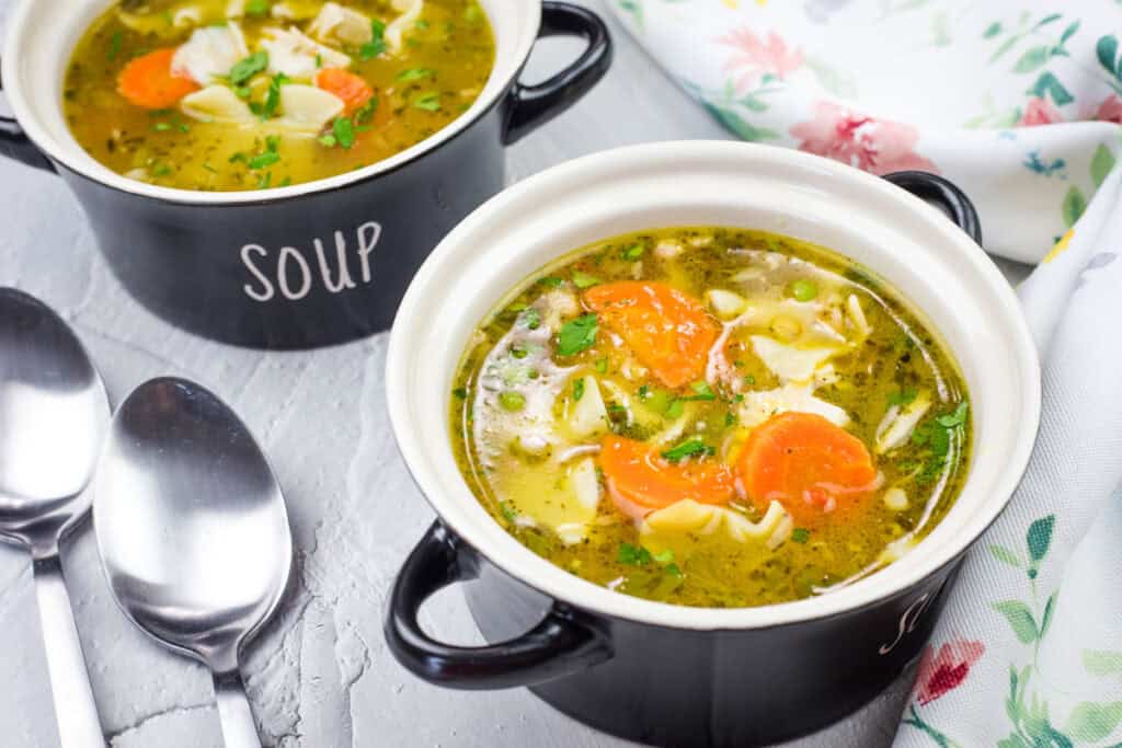Two black bowls labeled "SOUP" filled with chicken noodle soup, carrots, and herbs. Two silver spoons are placed beside them on a gray surface, with a floral cloth nearby.