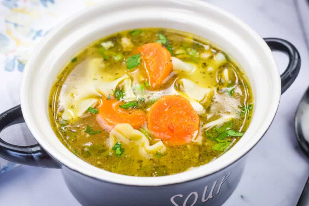 A black pot filled with chicken noodle soup, featuring carrots, parsley, and curly egg noodles in a clear broth.
