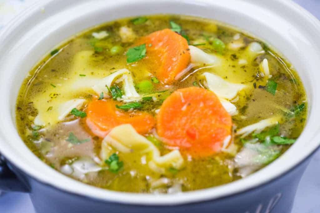 A bowl of chicken noodle soup with carrots, peas, and herbs in a broth.