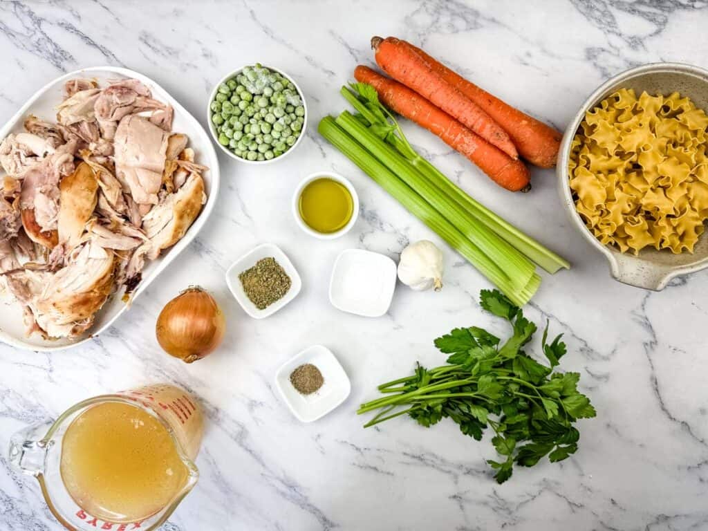 Ingredients on a marble countertop: shredded chicken, broth, onion, parsley, celery, carrots, frozen peas, garlic, egg noodles, olive oil, and seasonings in small bowls.