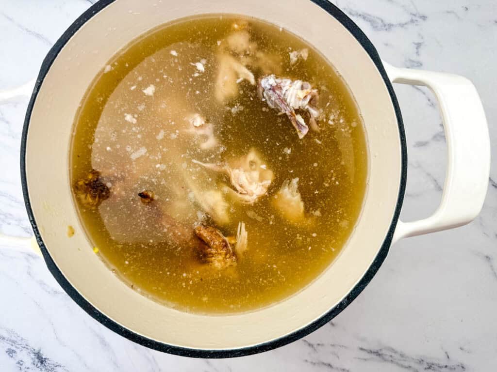 A pot of broth with visible chicken bones and pieces, on a white marble surface.