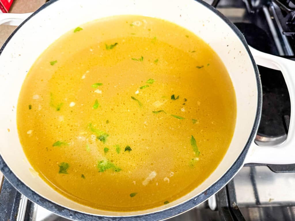 A pot of chicken broth with chopped herbs simmers on a stove.