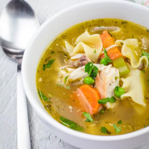 A bowl of chicken noodle soup with carrots, peas, celery, and herbs, next to a spoon on a textured white surface.