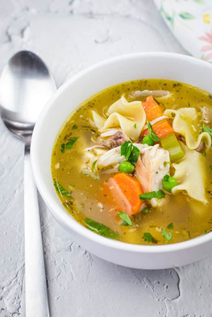 A bowl of chicken noodle soup with carrots, peas, celery, and herbs, next to a spoon on a textured white surface.