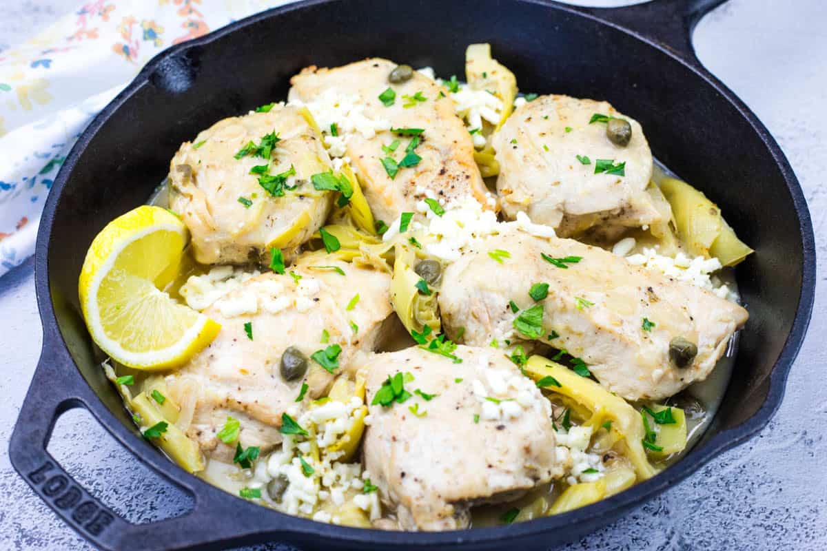 Low-carb skillet Chicken with Lemon, Artichokes & Feta in a black skillet, garnished with lemon wedges and parsley.