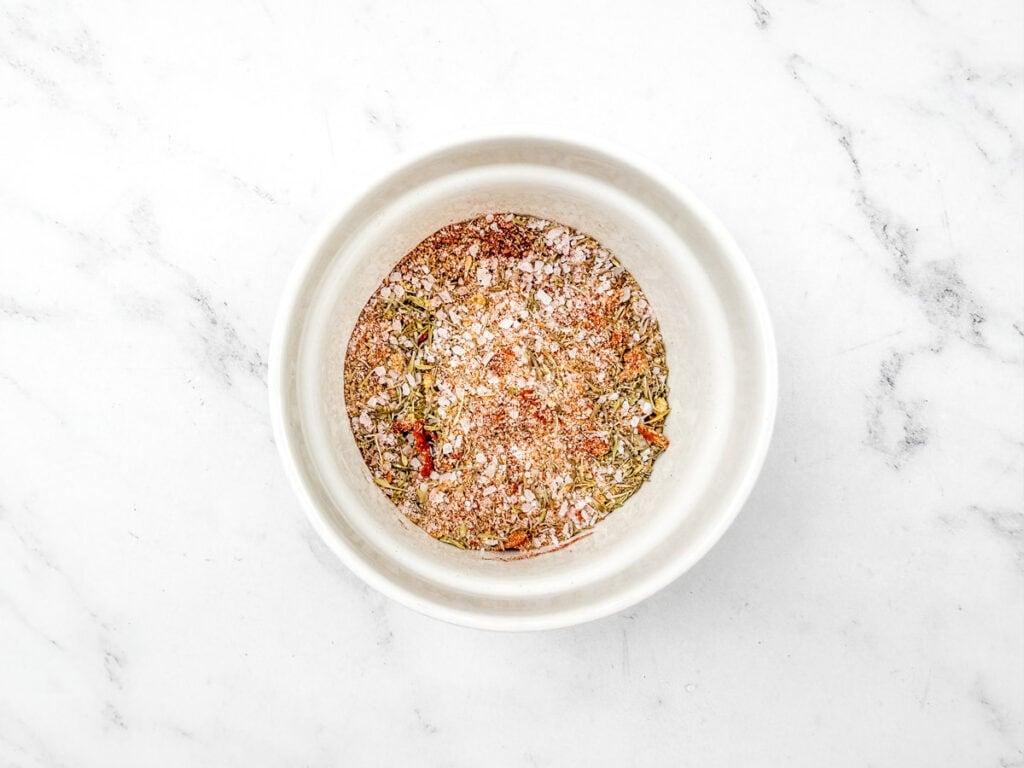 A combination of dried thyme, garlic powder, onion powder, paprika, salt, and a few grinds of freshly ground pepper in a small bowl on a white marble surface.