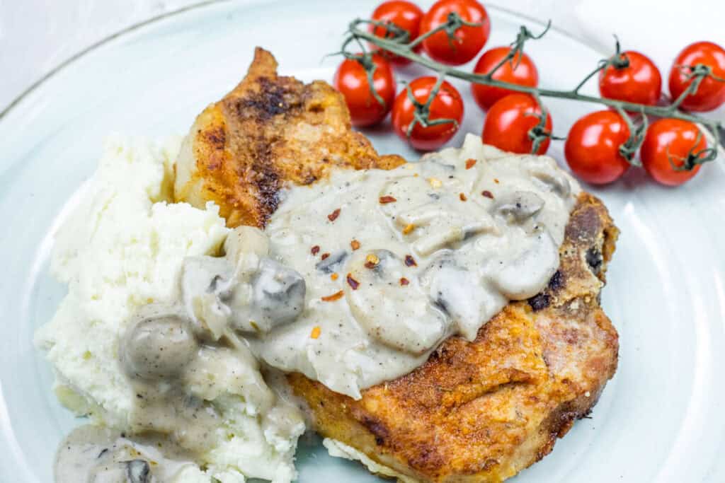 A plate with mashed potatoes, fried chicken topped with creamy mushroom sauce, and a bunch of cherry tomatoes on the side.