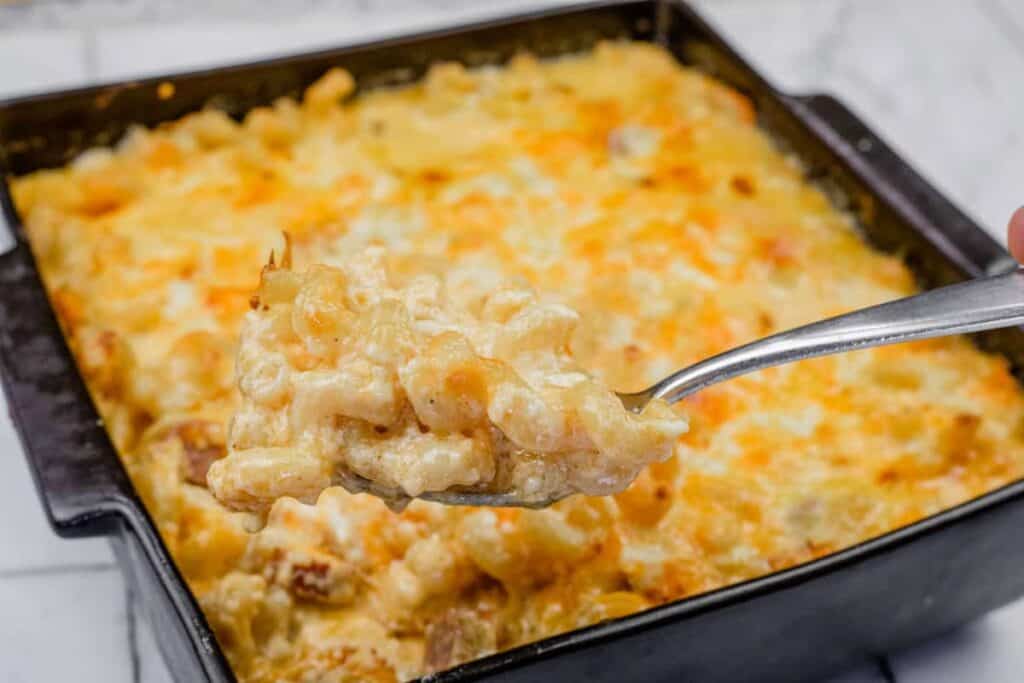 A spoonful of Tini&rsquo;s Mac & Cheese with a Twist is held above a square baking dish filled with the same dish.
