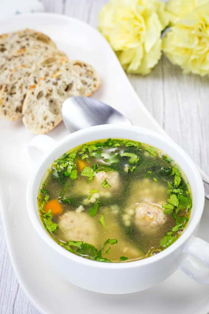 Italian wedding soup with meatballs, spinach, and pasta in a clear broth, served with sliced bread.